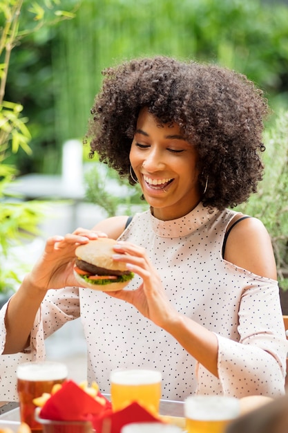 Vivaz feliz joven negra comiendo una hamburguesa sosteniéndola en sus manos con una sonrisa radiante y mirada de anticipación en un restaurante al aire libre