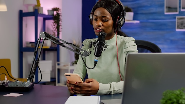 Vlogger africano gravando videoblog lendo mensagens de smartphone com equipamento moderno em podcast de estúdio