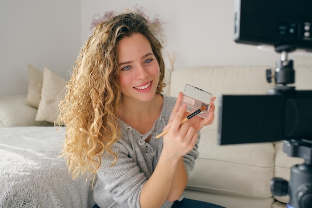 Vlogger de belleza sonriente con cabello rizado grabando tutorial de maquillaje cosmético mientras sostiene paleta y cepillo en casa