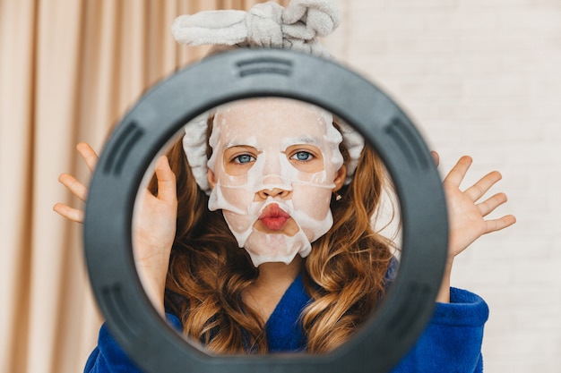 Foto vlogger de adolescente aplicando máscara facial de cuidados com a pele enquanto grava um vídeo na frente da lâmpada. garota feliz se divertindo na filmagem do tutorial usando um telefone móvel inteligente.