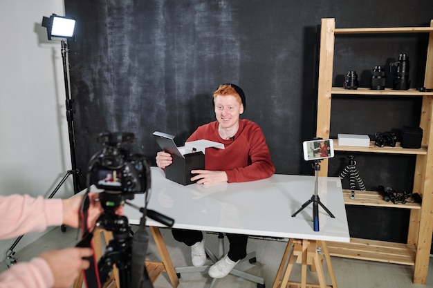 Vlogger masculino alegre por escritorio va a desempaquetar la caja con la nueva cámara fotográfica durante la grabación de video en el estudio