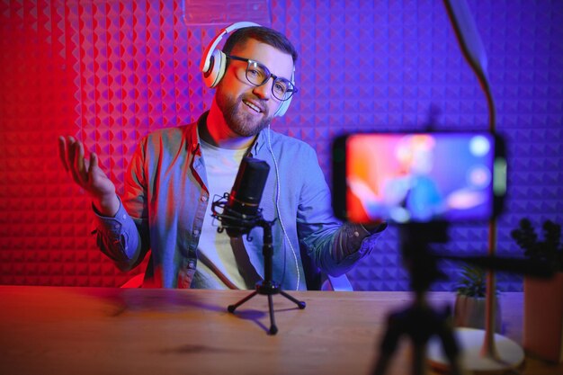 Vlogger que usa un teléfono inteligente para filmar un podcast en un blogger de estudio con un micrófono de teléfono móvil y auriculares que filman videos para una carrera de transmisión en las redes sociales