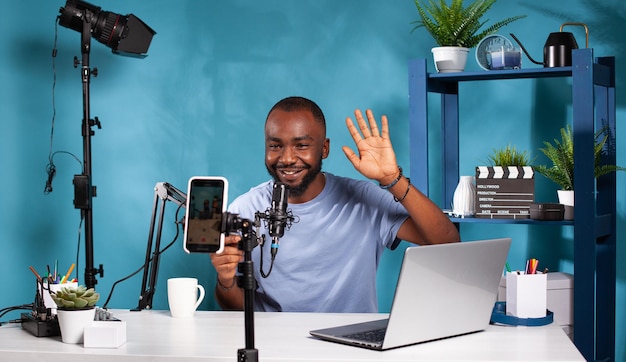 Vlogger sonriente saludando frente a un teléfono inteligente de grabación sentado en el escritorio con micrófono profesional. Creador de contenido haciendo gestos de saludo con la mano mirando la configuración de podcast de video en vivo.