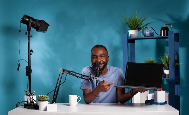 Vloguero afroamericano revisando un nuevo modelo de computadora portátil que señala características importantes sentadas en el escritorio con la configuración de podcasting. Influyente hablando en el micrófono del estudio sobre una computadora portátil.
