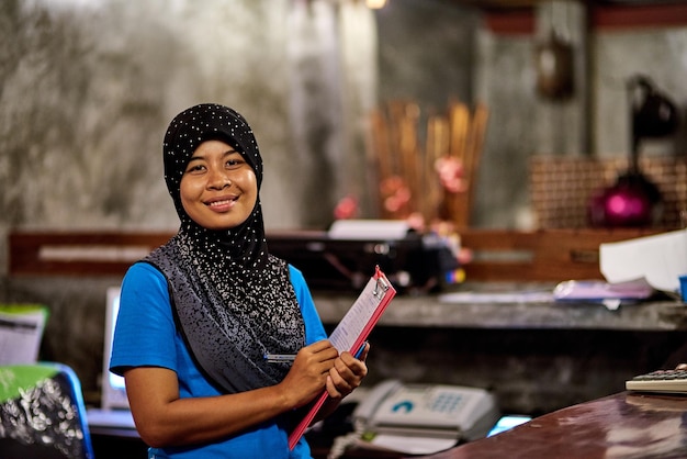 Você é sempre bem-vindo aqui Retrato de uma mulher tailandesa muçulmana sorridente em pé atrás de uma recepção de hotel segurando uma prancheta