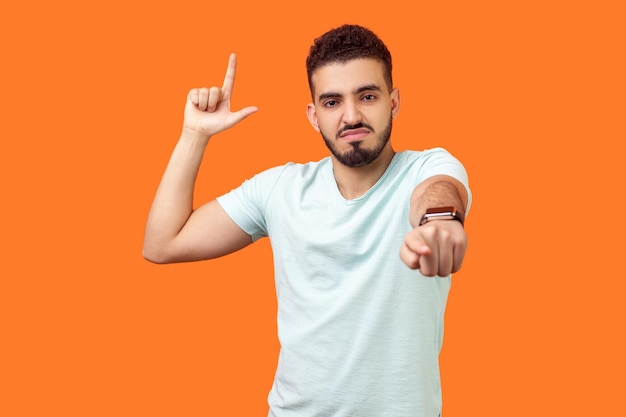 Você é um perdedor. Retrato de homem moreno zangado mandão com barba em camiseta branca, mostrando o gesto de perdedor e apontando para a câmera, zombando de sua derrota. tiro de estúdio interno isolado em fundo laranja