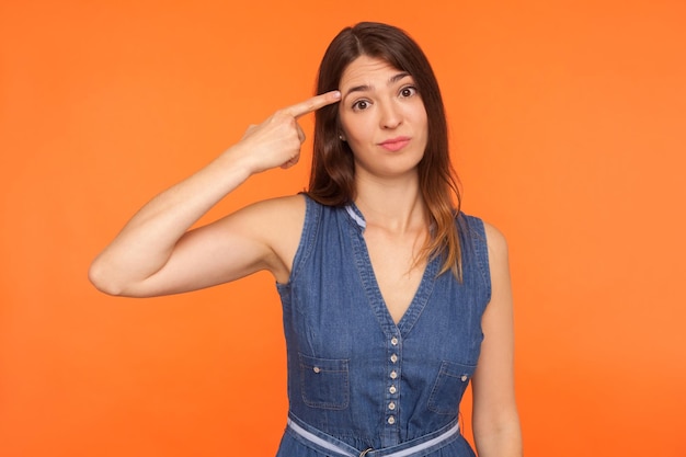 Você está brincando comigo Mulher morena positiva em vestido jeans segurando o dedo perto da têmpora da cabeça e gesticulando Você é um idiota estúpido parecendo descontente com a ideia maluca estúdio interno tiro fundo laranja