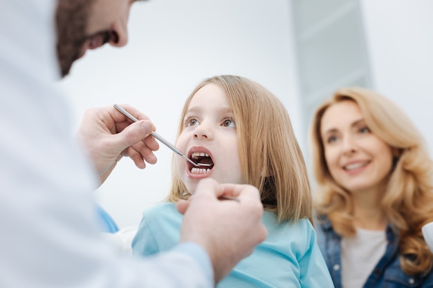 Você não é assustador. admirável garota incrível ativa sentada na cadeira enquanto um dentista olha para os dentes dela usando um equipamento especial