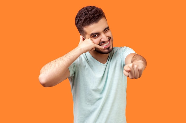 Você nos contata retrato de homem moreno positivo com barba em camiseta branca fazendo gesto com os dedos discar meu número ou me ligar de volta e apontando para você tiro de estúdio isolado em fundo laranja