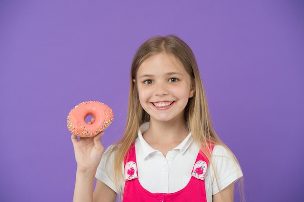 Você quer morder O rosto sorridente de menina segura rosquinha rosa na mão fundo violeta Garota sorridente pronta para morder rosquinha Conceito de lanche A criança não pode esperar para comer rosquinha doce espaço de cópia