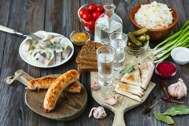 Vodka con manteca de cerdo, pescado salado y verduras, salchichas sobre superficie de madera. Bebida artesanal pura de alcohol y merienda tradicional, tomates, repollo, pepinos. Espacio negativo. Celebrando la comida y deliciosa.