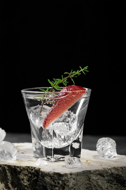 Vodka o ginebra en vaso de chupito con hielo decorado con rodaja de fresa y tomillo en podio de travertino