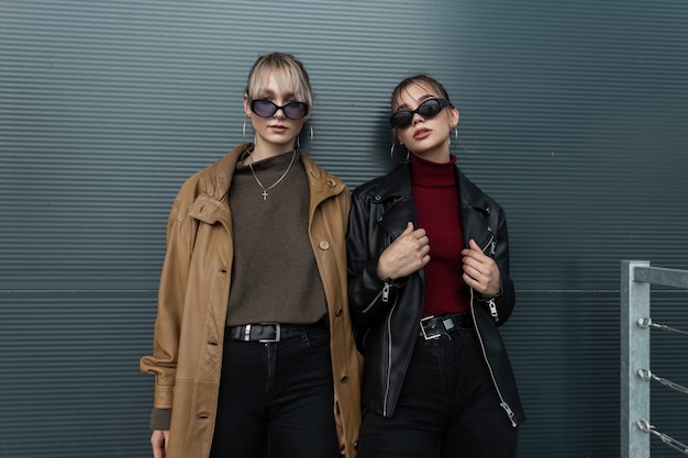 Vogue duas mulheres elegantes com óculos escuros estilosos, jaquetas de couro e jeans preto posando perto de uma parede de metal na rua