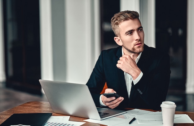 Volle Konzentration. Ein junger Geschäftsmann sitzt mit einem Laptop in einem Business Center, legt seine Hand mit dem Telefon in der Hand auf sein Kinn, auf dem Tisch liegen Dokumente und Kaffee