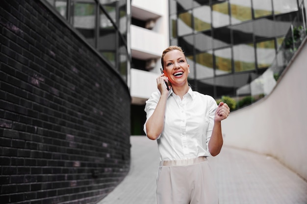 Volle Länge der attraktiven blonden modischen Geschäftsfrau, die am Telefon im Außenbereich des Geschäftszentrums spricht.