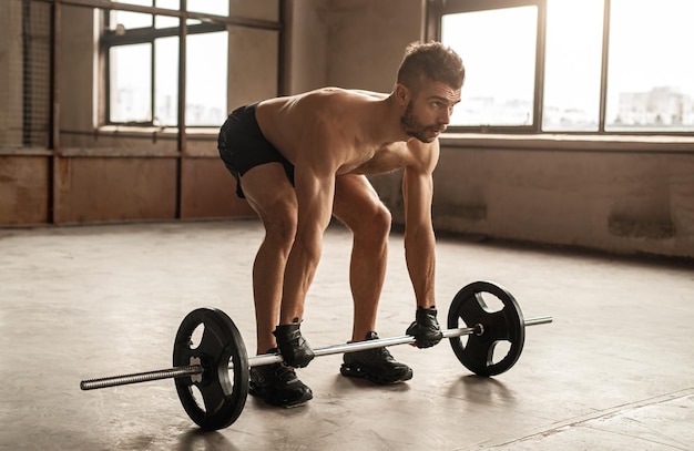 Volle Länge des konzentrierten athletischen, hemdlosen, bärtigen Mannes in Gewichtheberhandschuhen, der während des intensiven Trainings im Fitnessstudio schwere Langhantel hebt