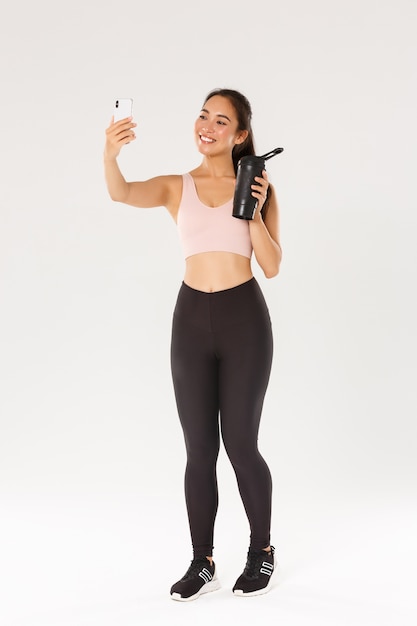 Volle Länge des lächelnden schlanken und gesunden asiatischen Mädchens, das Selfie-Fitness-Trainingssitzung nimmt, Bloggerin und Athelte, die Wasserflasche auf Kamera des Handys zeigt, über weißem Hintergrund stehend.