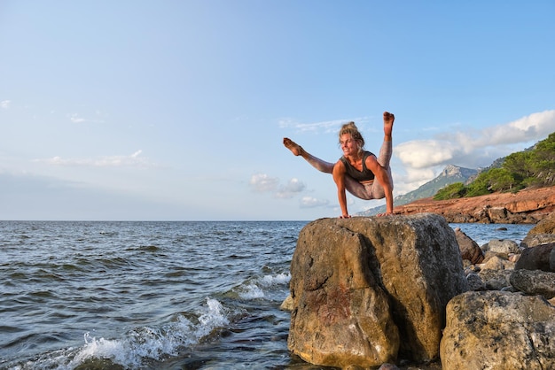 Volle Länge einer lächelnden Sportlerin in Sportkleidung, die in Firefly-Pose auf einem Felsbrocken balanciert und das plätschernde Meer bewundert, während sie bei Sonnenuntergang Yoga praktiziert