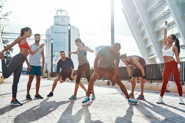 Volle Länge von Menschen in Sportkleidung, die sich beim Training auf dem Bürgersteig im Freien aufwärmen und dehnen