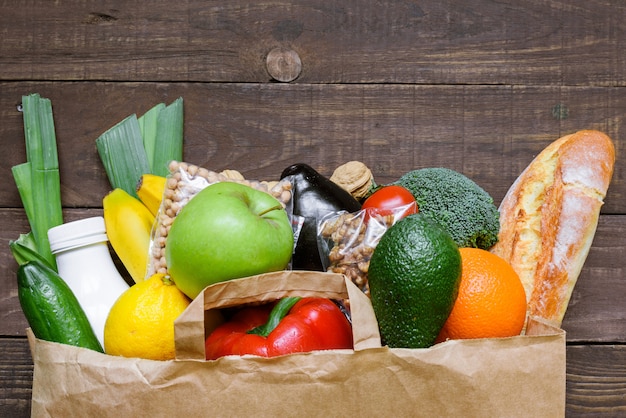 Volle Papiertüte mit verschiedenen gesunden vegetarischen Lebensmitteln auf rustikalem Holztisch. Draufsicht