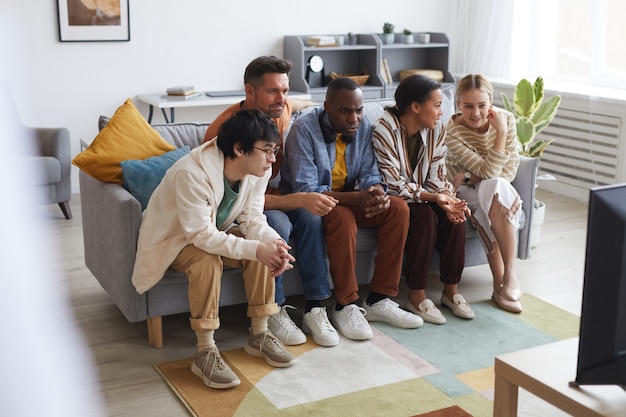 Voller Blick auf eine multiethnische Gruppe von Freunden, die zu Hause zusammen fernsehen, während sie in einer Reihe auf dem Sofa sitzen, Platz kopieren