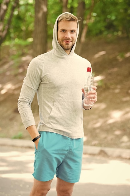 Voller Energie Mann in Kapuze trinkt Wasser erfrischendes Vitamingetränk nach dem Training sportlicher Mann mit Wasserflasche Athlet trinkt Wasser nach dem Training im Park Pflege Körper Hydratation Sport und Gesundheit