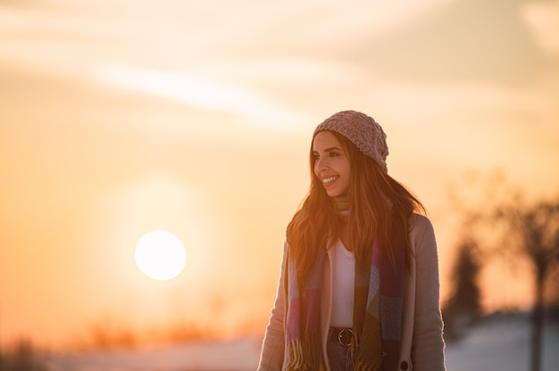 Voller Körper der glücklichen jungen Frau in der warmen Kleidung, die auf frischem weißem Schnee in der Winterlandschaft zur Sonnenuntergangszeit spaziert