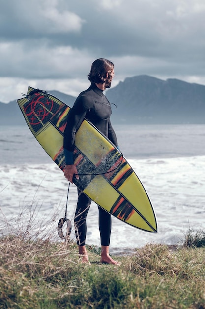 Voller Körper eines jungen, muskulösen, bärtigen männlichen Athleten im schwarzen Neoprenanzug mit Surfbrett in der Hand, der an der grasbewachsenen Küste steht und wegschaut, nachdem er im Meer vor bewölktem Himmel das Surfen geübt hat