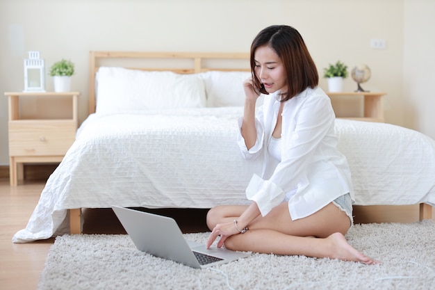 Voller Schuss der erwachsenen freiberuflich tätigen asiatischen Frau im weißen Hemd, das an Computer und Handy im Schlafzimmer arbeitet