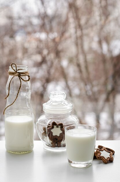 Volles Glas Milch, Schokoladenkekse und eine Flasche auf Frühstückstisch auf Fensteransicht.