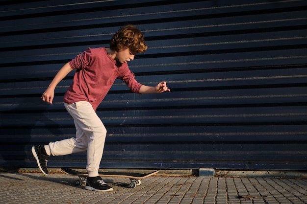 Vollkörperiger Junge in lässiger Kleidung, der beim Skateboardfahren gegen eine dunkelblaue Wand auf einer Stadtstraße rennt