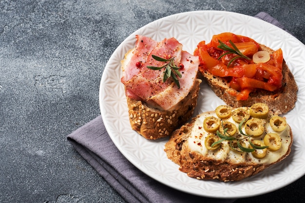 Vollkornbrot-Sandwiches mit Frischkäse, Speck und Oliven in Dosen Paprika mit Tomaten auf einem Teller.