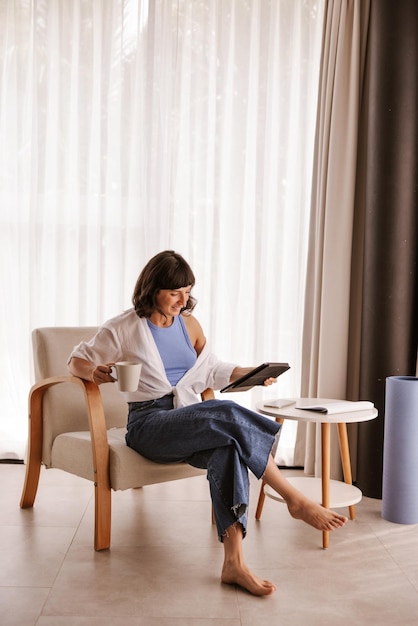 Vollständige ansicht einer frau, die auf einem stuhl sitzt und auf einen laptop blickt, der eine tasse kaffee hält