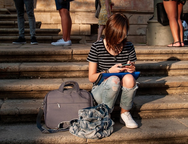 Foto vollständige länge von teenager mit mobiltelefon, während sie auf den stufen sitzt
