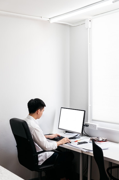 Volta do homem vestindo camisa branca e sentar na cadeira de escritório preta, trabalhando com seu computador pessoal com display na mesa do escritório.