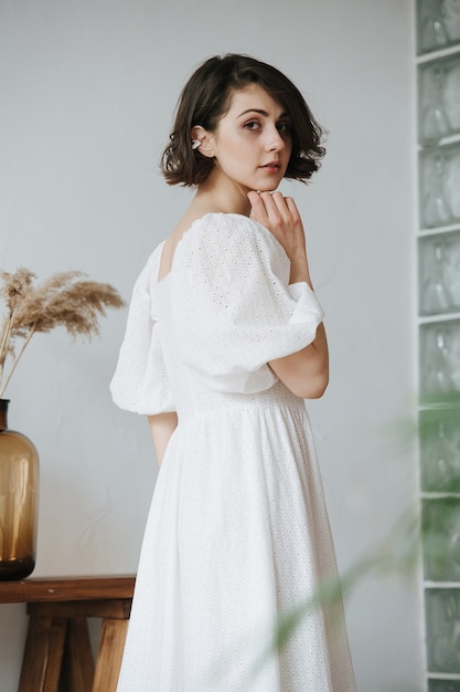 Voltando a mulher tímida sensual em um vestido branco. Bela morena com cabelo curto dentro de casa.