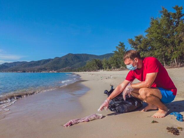 Voluntario activista Hombre con mascarilla médica recoge basura que contamina la arena de la playa cerca del mar Problema de basura derramada causada por la contaminación del planeta hecha por el hombre y el concepto de protección ambiental