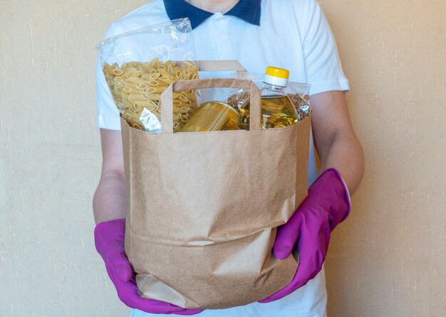 Voluntario en los guantes protectores poniendo comida en caja de donación. Repartidor empleado en chaleco rojo caja de embalaje con comida. Servicio de cuarentena de pandemia de coronavirus.