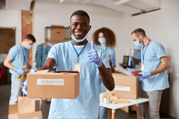 Voluntario masculino joven afroamericano en máscara protectora uniforme azul y guantes sonriendo mostrando