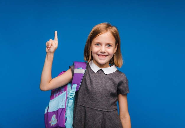 Volver a la escuela linda colegiala positiva en uniforme levantó el dedo posando sobre un fondo azul Preparación para la escuela una idea interesante Espacio para copiar