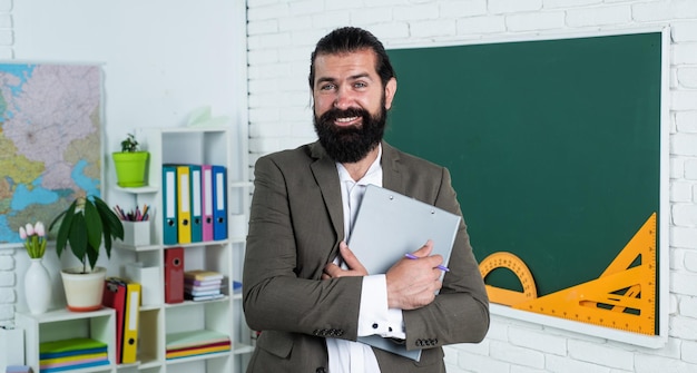 Volviendo a la clase, un hombre feliz con barba sosteniendo un cuaderno para estudiar a un estudiante de educación informal en el aula de la escuela mientras la lección aprueba el examen aprendiendo el tema