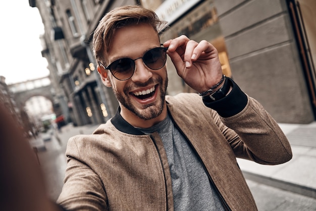 Volviéndose loco. Autorretrato de un joven apuesto en ropa casual sonriendo y ajustando sus gafas mientras está de pie al aire libre