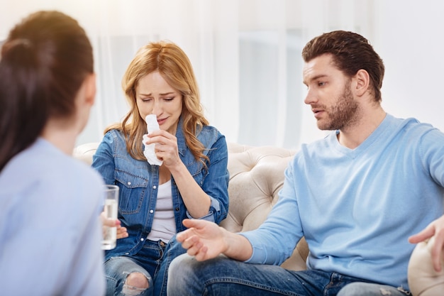 Von Emotionen überwältigt. Unglückliche traurige junge Frau wischte sich die Nase mit einem Papiertaschentuch ab und nahm ein Glas Wasser, während sie dem Psychologen von ihren familiären Problemen erzählte