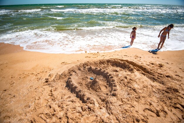 Von Kindern gebaute Sandfestung steht inmitten von zwei kleinen Mädchen, die von lauten Meereswellen am Strand stehen