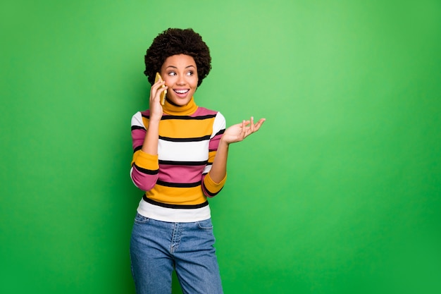 von lustigen hübschen dunklen Haut wellige Dame hält Telefon in der Nähe von Ohr sprechen besten Freund hören frische Klatsch Gerüchte tragen lässig gestreifte Pullover Jeans