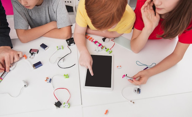 Von oben aus nicht erkennbare Kinder mit Lehrer, die sich am Tisch versammeln, mit Tablet und kleinen elektronischen Details, während sie während des Technologieunterrichts in der Schule Roboter zusammen bauen