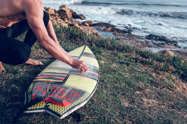 Von oben ein Surfer mit nacktem Oberkörper, der sein Surfbrett mit Wachs bedeckt und auf einer Hocke an der grasbewachsenen Küste sitzt