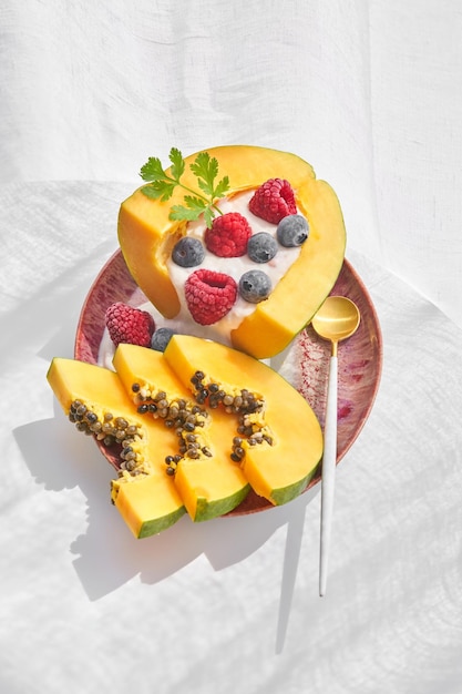 Von oben eine Portion süßes Papaya-Dessert, serviert auf einem Keramikteller mit Löffel und dekoriert mit Joghurt, Himbeere und Blaubeeren auf weißem Hintergrund