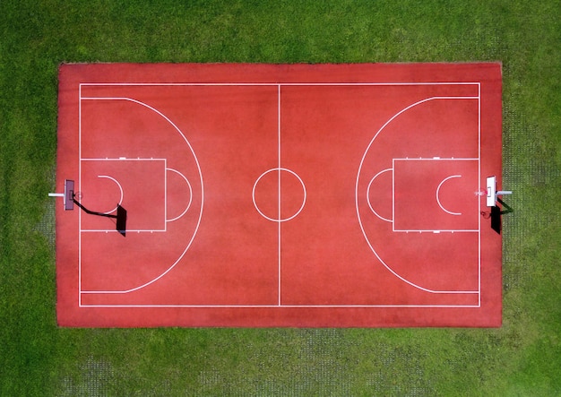 Von oben Luftaufnahme des leeren roten Basketball-Sportplatzes mit weißen Markierungen und Reifen, umgeben von grünem Gras