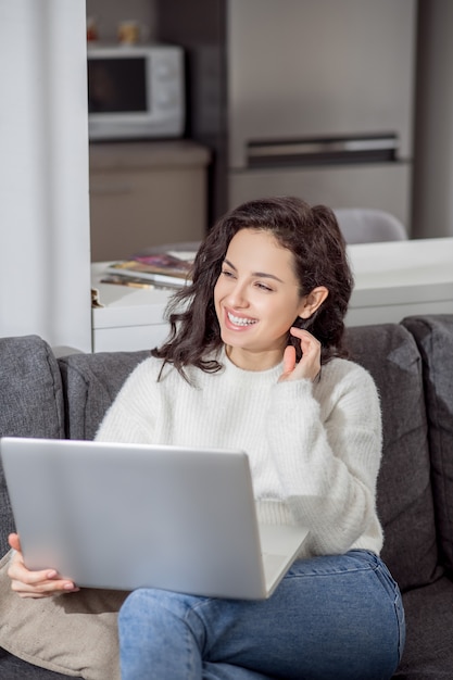 Von zuhause aus arbeiten. Eine junge Frau in einem weißen Pullover, der mit einem Laptop auf dem Sofa sitzt